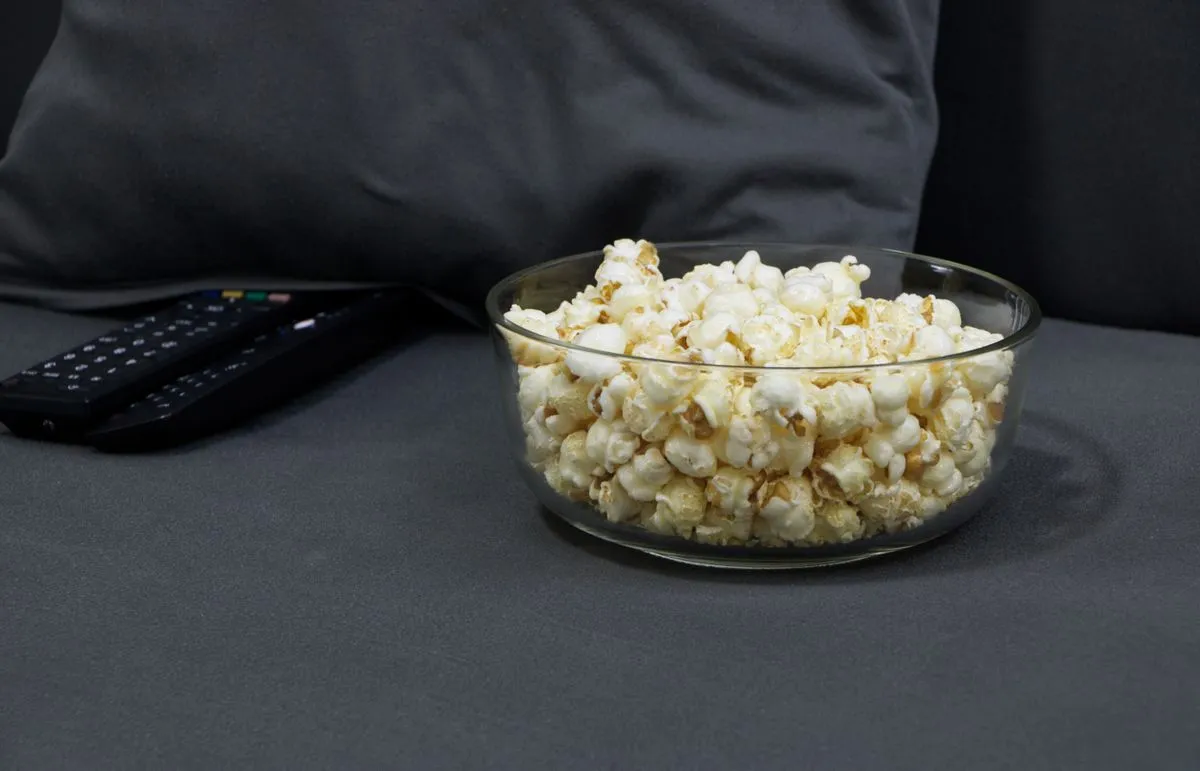 Bowl of popcorn next to TV remotes on a grey sofa
